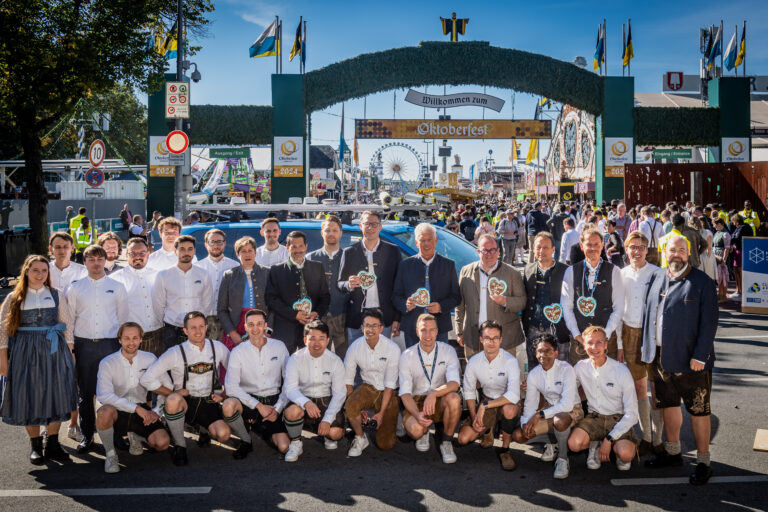 Gruppenfotos des MCube Wiesn Shuttle Teams: Erste Reihe kniend, zweite Reihe stehend. Alle tragen Tracht, sechs Personen haben Lebkuchenherzen um den Hals. Das Foto wurde vor dem Haupteingang des Oktoberfests aufgenommen.