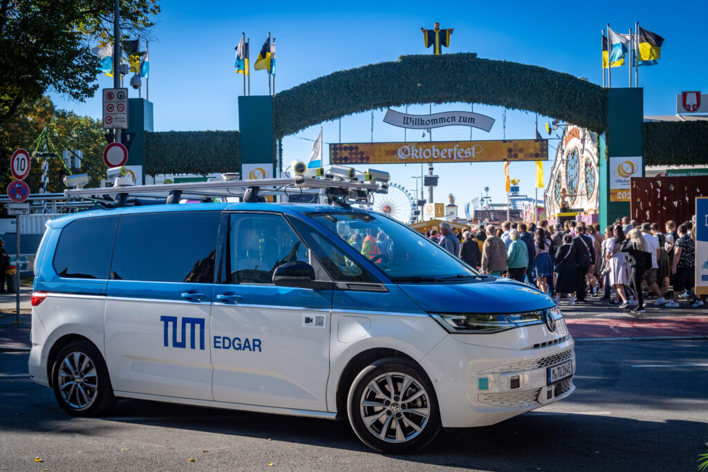 Ein autonom fahrender Volkswagen-Bus mit der Beschriftung „TUM EDGAR“ steht vor dem Eingang des Oktoberfests in München. Im Hintergrund sind viele Besucher, bayerische Fahnen und das festliche Eingangstor zu sehen. Das Fahrzeug ist mit Sensoren und Kameras ausgestattet.