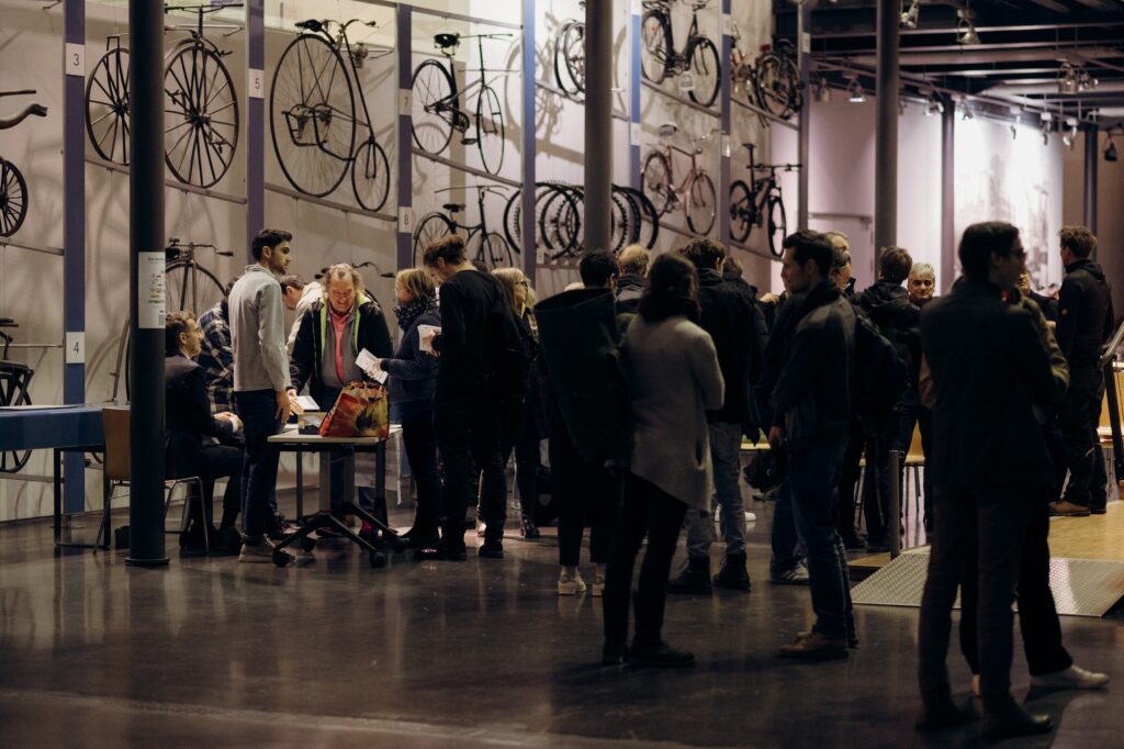 Eine Gruppe von Menschen steht und unterhält sich in einem Museum mit einer Ausstellung historischer Fahrräder an den Wänden. Die Atmosphäre wirkt lebendig und gesellig, mit Besuchern unterschiedlichen Alters.