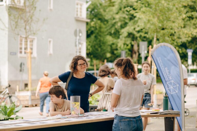 Eine Mutter und ihr Sohn stehen vor einem MCube-Informationsstand und erhalten Auskünfte von einer Mitarbeiterin.