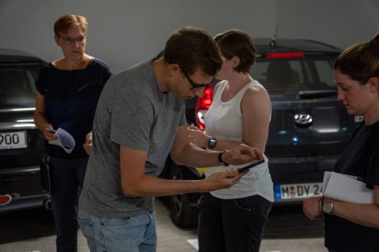 Vier Personen stehen in einer Tiefgarage zwischen geparkten Autos. Ein junger Mann im grauen T-Shirt zeigt einer Frau auf seinem Smartphone etwas. Die übrigen Personen beobachten die Szene aufmerksam. Zwei der Personen halten Blätter oder Notizen in der Hand.