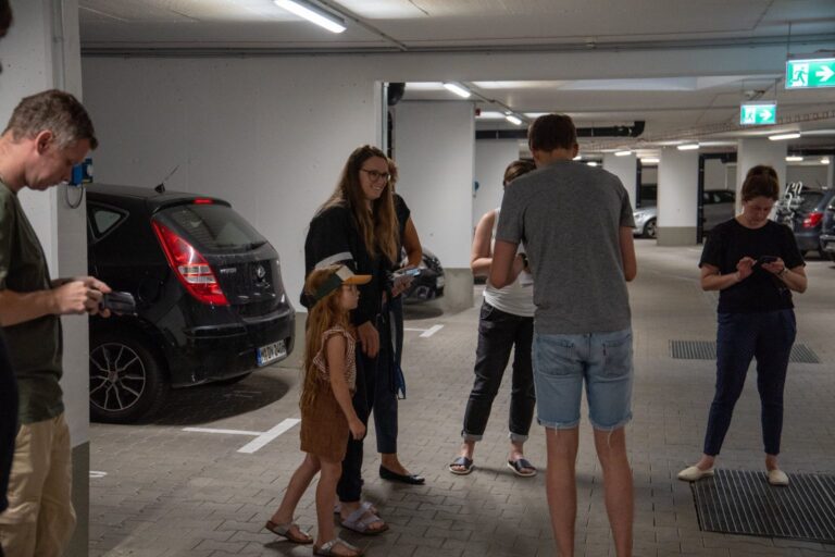 Mehrere Personen, darunter ein Kind, befinden sich in einer Tiefgarage zwischen geparkten Autos. Eine Frau lächelt, während sie neben dem Kind steht. Einige Personen schauen auf ihre Smartphones, andere halten Geräte oder Zettel in der Hand. Die Szene wirkt wie Teil einer Führung oder interaktiven Aktivität.
