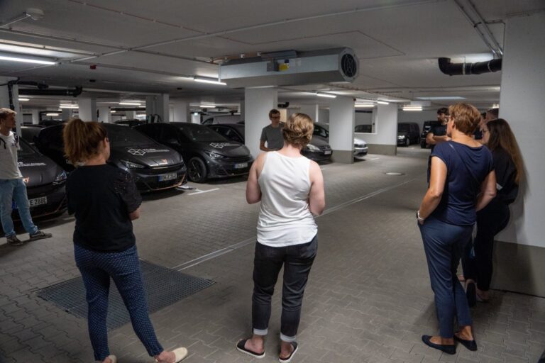 Eine Gruppe von Menschen steht in einer Tiefgarage und hört einer Person zu, die vor einer Reihe schwarzer Fahrzeuge spricht. Die Fahrzeuge sind mit Logos und Texten beschriftet, vermutlich im Zusammenhang mit einem Forschungs- oder Mobilitätsprojekt. Die Szene wirkt wie eine Führung oder Präsentation.