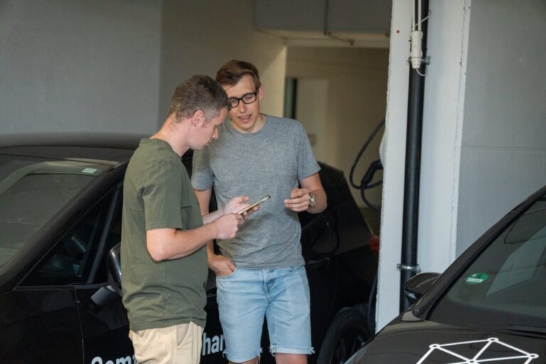 Zwei junge Männer stehen nebeneinander in einer Tiefgarage und schauen gemeinsam auf ein Smartphone. Einer von ihnen trägt ein graues T-Shirt und Jeansshorts, der andere ein grünes T-Shirt und beige Hosen. Im Hintergrund stehen schwarze Autos mit Beschriftung, möglicherweise Teil eines Carsharing- oder Forschungsprojekts.
