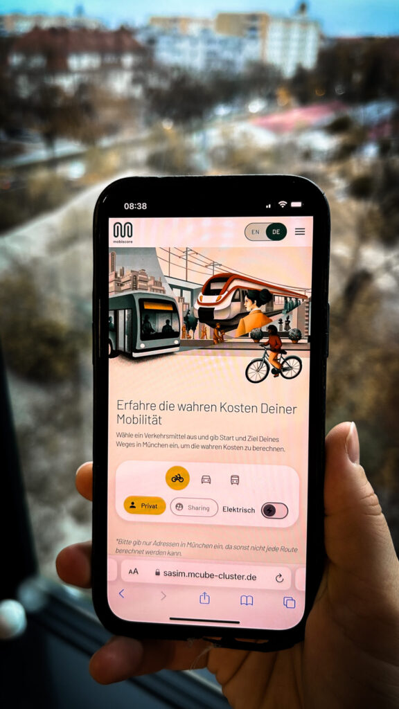 Foto einer Hand, die ein Smartphone hält. Auf dem Display ist die Webseite „sasim.mcube-cluster.de“ geöffnet, die die wahren Kosten der Mobilität in München berechnet. Die Seite zeigt Auswahloptionen für verschiedene Verkehrsmittel wie Fahrrad, Auto und öffentliche Verkehrsmittel sowie Einstellungen für privat, Sharing und elektrisch. Im Hintergrund ist eine unscharfe Stadtansicht zu erkennen.