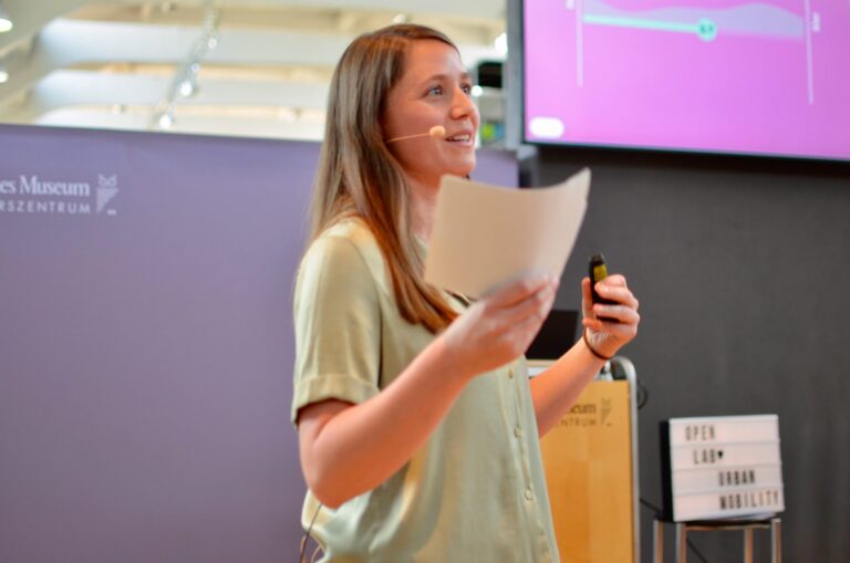 Eine Frau steht vor einem Rednerpult im Deutschen Museum Verkehrszentrum und hält ein Blatt Papier in der Hand. Sie spricht mit einem Headset-Mikrofon und hält einen Präsentationsklicker. Neben ihr steht ein beleuchteter Kasten mit der Aufschrift ‚OPEN LAB URBAN MOBILITY