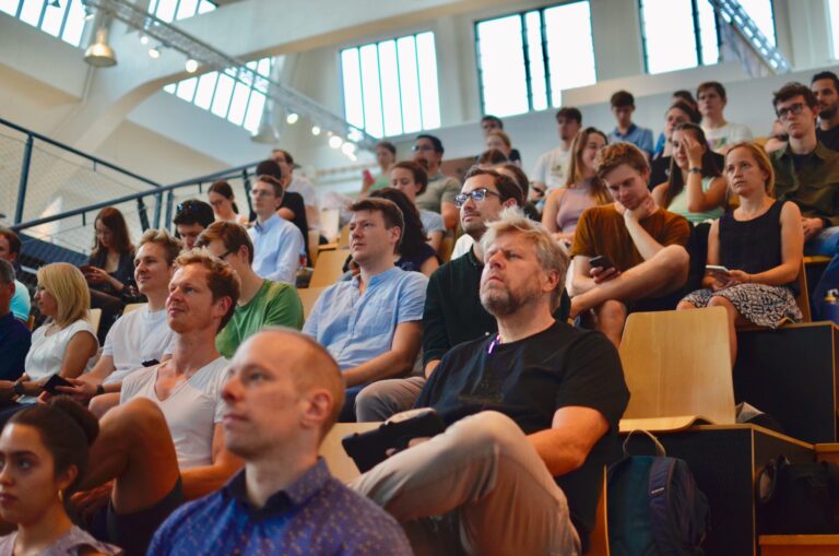 Zuschauerinnen und Zuschauer sitzen in einem hellen Auditorium auf Holzstühlen. Die meisten schauen aufmerksam nach vorne, einige haben Smartphones in der Hand. Die Gruppe ist vielfältig und konzentriert sich auf das Geschehen vor ihnen
