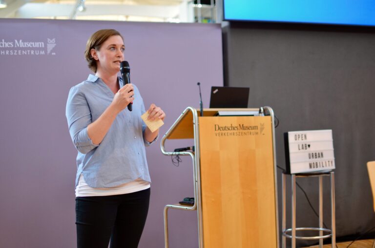 Eine Frau steht vor einem Rednerpult im Deutschen Museum Verkehrszentrum und spricht in ein Mikrofon. Sie hält einen kleinen Zettel in der Hand. Neben dem Pult steht ein beleuchteter Kasten mit der Aufschrift ‚OPEN LAB URBAN MOBILITY