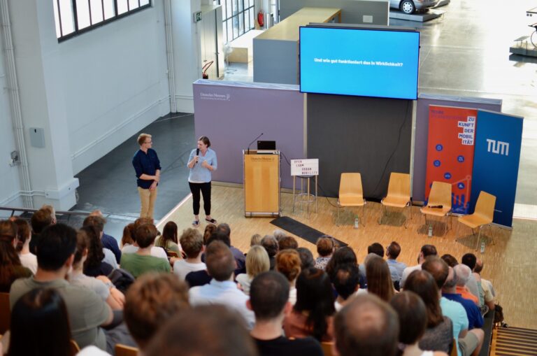In einem hellen Auditorium stehen zwei Personen vor einem Publikum. Die Frau spricht in ein Mikrofon, der Mann hört zu. Im Hintergrund sind ein Rednerpult mit dem Schriftzug ‚Deutsches Museum Verkehrszentrum‘, ein beleuchteter Kasten mit ‚OPEN LAB URBAN MOBILITY‘ und mehrere leere Stühle zu sehen. Auf einem großen Bildschirm ist die Frage ‚Und wie gut funktioniert das in Wirklichkeit?‘ zu lesen