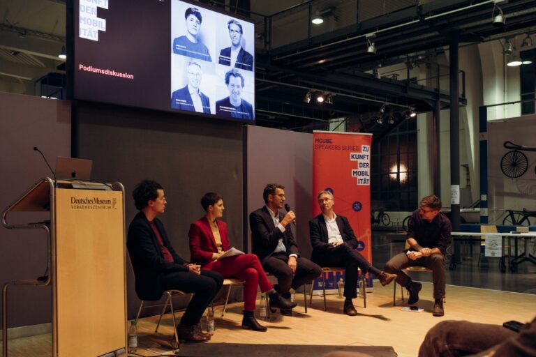 Auf diesem Bild ist eine Podiumsdiskussion zu sehen, im Deutschen Museum Verkehrszentrum, wie der Schriftzug am Rednerpult nahelegt. Es sitzen fünf Personen auf Stühlen auf der Bühne, vier Männer und eine Frau in der Mitte in rotem Anzug. Der Mann in der Mitte spricht gerade in ein Mikrofon. Im Hintergrund sind zwei Banner zu sehen, einer von der Technischen Universität München (TUM) und ein anderer mit dem Titel „MCube Speakers Series: Zukunft der Mobilität“.