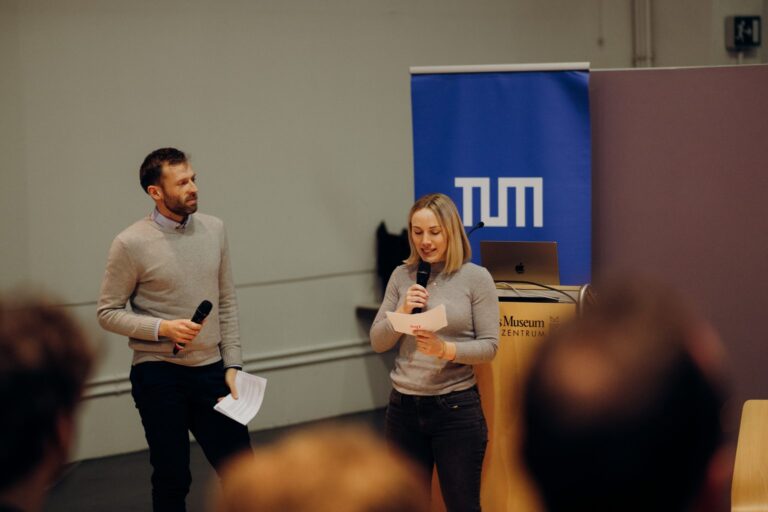 Das Bild zeigt zwei Personen, einen Mann und eine Frau, die vor einem Publikum sprechen. Beide halten Mikrofone und die Frau liest gerade von einem Blatt Papier ab. Im Hintergrund sieht man einen blauen Banner mit dem Logo der Technischen Universität München (TUM) und einen Rednerpult mit der Aufschrift "Deutsches Museum Verkehrszentrum". Das Setting sieht aus wie eine formelle Veranstaltung oder Präsentation