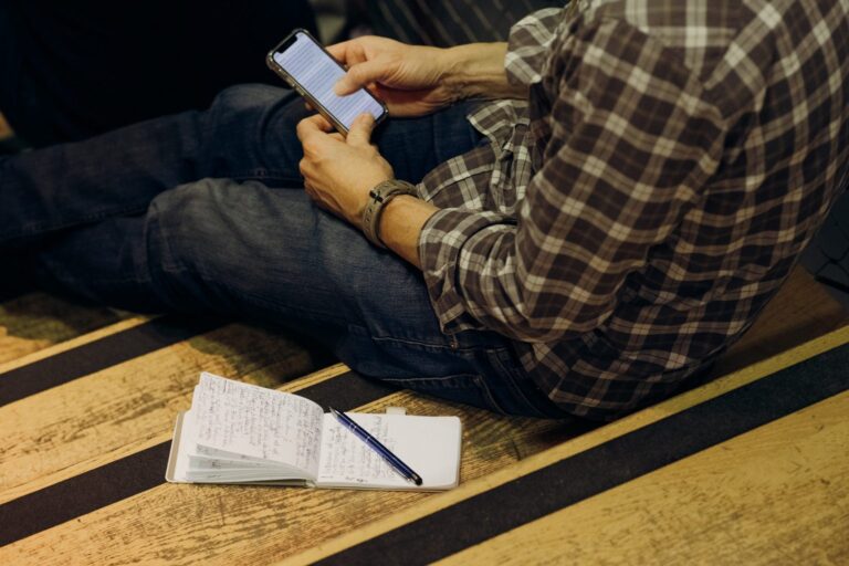 Das Bild zeigt eine Person, die auf einer Holztreppe sitzt. Sie trägt eine karierte Hemd und Jeans und hält ein Smartphone in den Händen, auf dem sie offenbar Text liest. Vor der Person liegt ein geöffnetes Notizbuch mit handschriftlichen Notizen und einem Stift darauf.