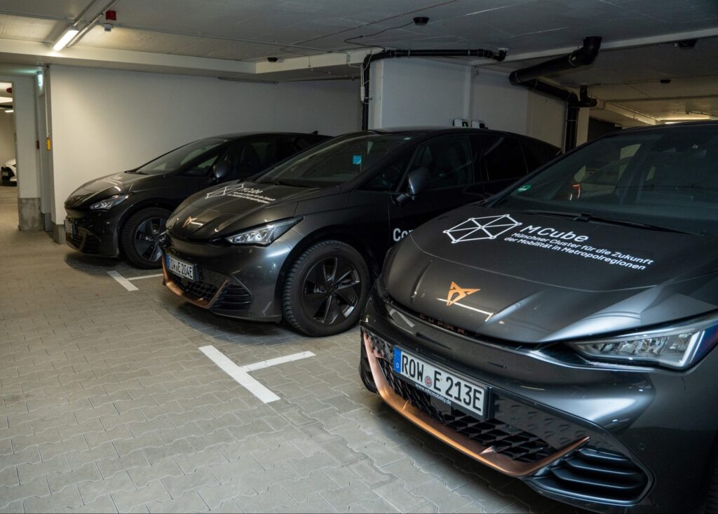 Das Bild zeigt drei Elektroautos, die in einer Tiefgarage nebeneinander geparkt sind. Alle Fahrzeuge sind dunkelgrau und tragen Logos von MCube, dem Münchner Cluster für die Zukunft der Mobilität in Metropolregionen, auf der Motorhaube. Die Wagen sind modern und scheinen zur Forschung oder zum Testen im Bereich nachhaltiger Mobilität eingesetzt zu werden.