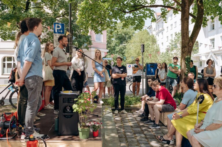 Seitlicher Blick auf die Bühne mit Mitarbeitenden des Projektes aqt und auf die ersten Reihen des Publikums bei der Eröffunungsrede des Sommerfestes der Südlichen Au.