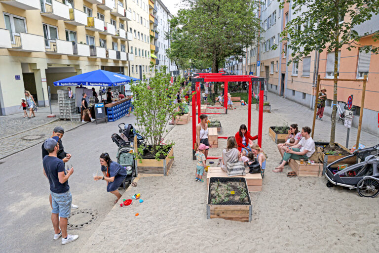 Blick auf die umgestaltete Straße. Ein Teil der Straße wurde in einen Sandkasten umgewandelt, der von Familien mit jungen Kindern sehr gut angenommen wird.