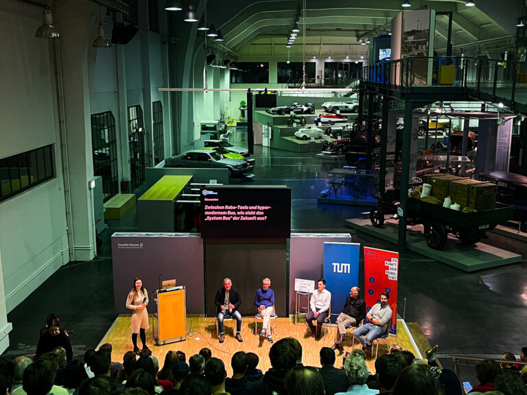 Blick von der Tribüne im Verkehrszentrum des Deutschen Musuems (Halle 3), mit Blick auf die Teilnehmenden der Podiumsdiskussion der Speaker Series #7 mit dem Thema "Das System Bus radikal neu gedacht" und Autos in der Museumshalle im Hintergrund.