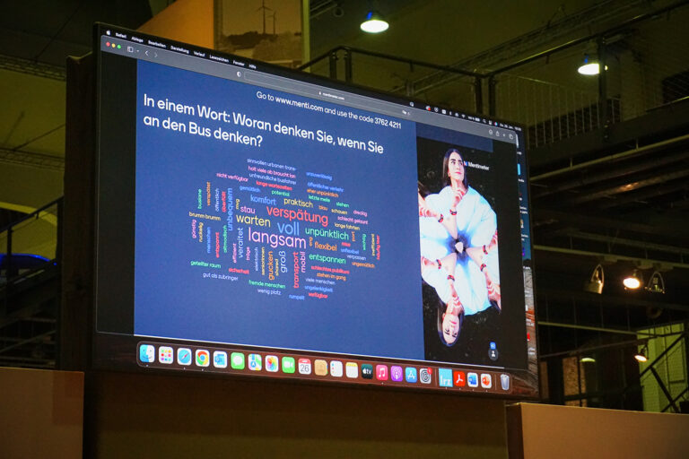 Blick auf die Bühne im Verkehrszentrum des Deutschen Musuems (Halle 3) auf die Leinwand, die eine Folie der Speaker Series #7 mit dem Thema "Das System Bus radikal neu gedacht" zeigt.