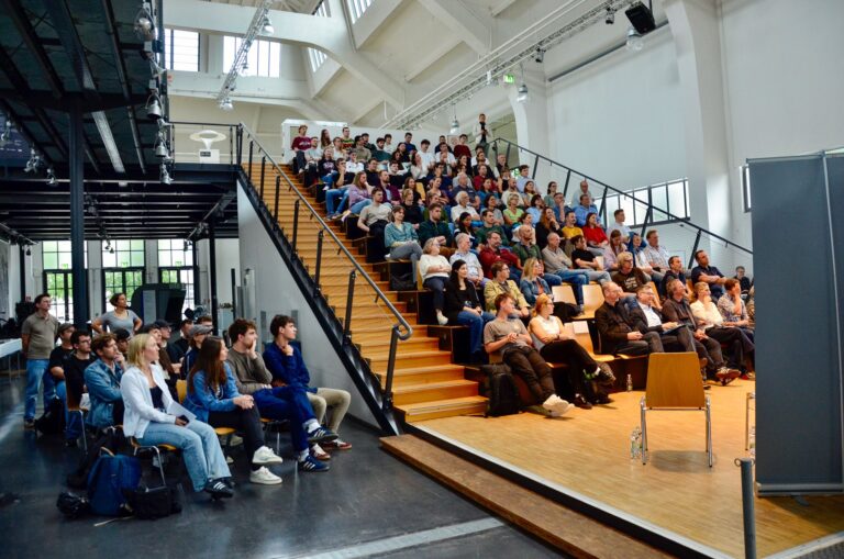 Vollbesetzte Halle 3 im Verkehrszentrum des Deutschen Museums während der MCube Speaker Series #20 mit dem Thema "Olympia als Entwicklungsmotor?", Blick von der Bühne ins Publikum.