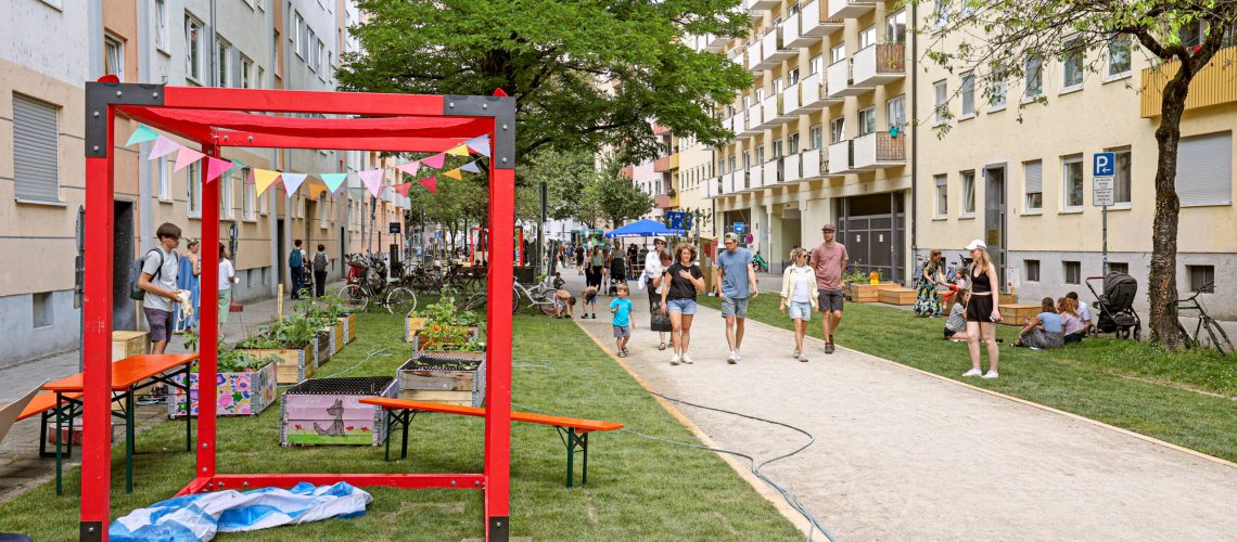 Blick auf die umgestaltete Straße, die statt Parkplätzen nun Grünflächen und einen Fußgänger- und Radfahrweg in der Mitte hat. Die Grünflächen und Wege werden von den Teilnehmenden des Sommerfestes gerne benutzt.